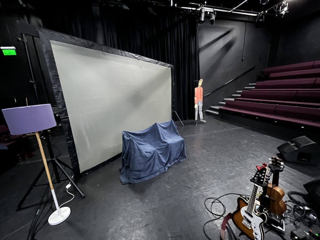The stage suring set up. A large blank back-projection screen hangs across the width of the stage with a standard lamp on the lefthand side of it. In the centre of the stage two chairs are covered with a fleecy blankeys to approximate the appearance of a sofa. to the right rows of upholstered bench seats are visible