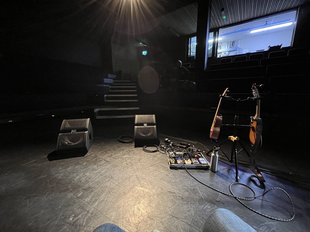 view from the stage. Padded bench seats are just visible in the darkness to the left and right, with steps up straight ahead. Two speakers sit a little forward of the steps, one on each side. to the right of them is an effects peddle board and a stand holding a ukulele and an electric mandolin